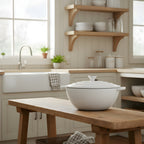 White casserole dish on a wooden table in a kitchen with shelves and windows in the background.